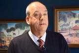 A bald, older man wearing a pullover and tie stands in front of some paintings.