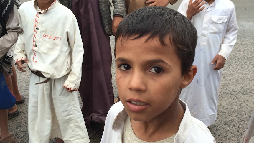 A group of kids in Yemen's Amran province pose for a photo. They are standing by a roadside, there is some grass but no trees.