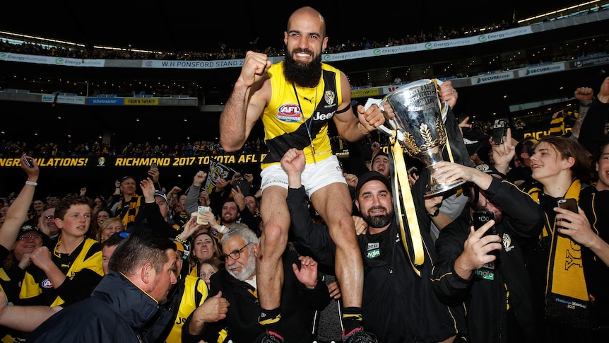 Bachar Houli stands on the MCG fence holding the AFL premiership cup. A crowd of Tigers fans celebrates behind him