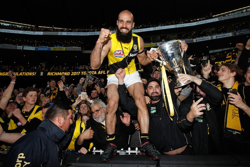 Bachar Houli stands on the MCG fence holding the AFL premiership cup. A crowd of Tigers fans celebrates behind him