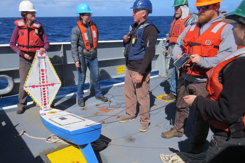 Six people standing on a large boat out in the ocean preparing to launch a mini boat