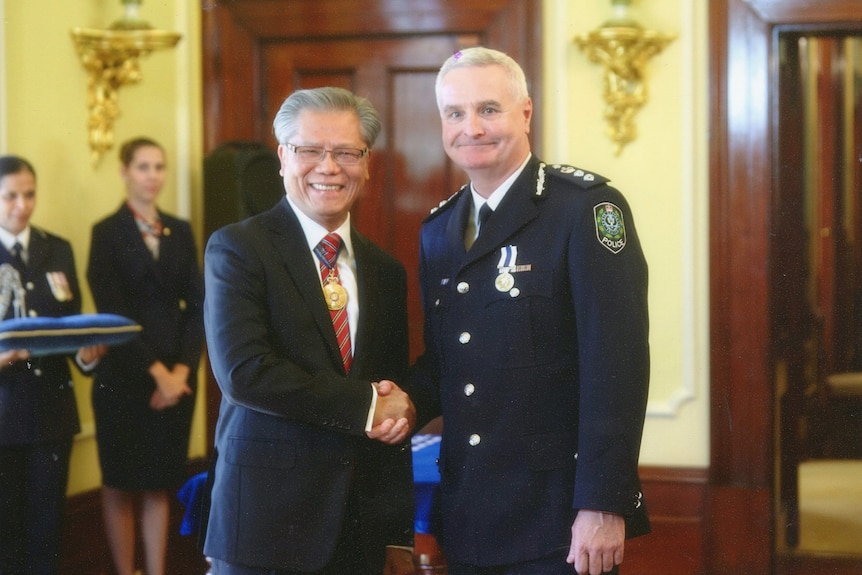 A man with grey hair and a suit shakes the hand of another, taller man with grey hair, wearing a formal police uniform