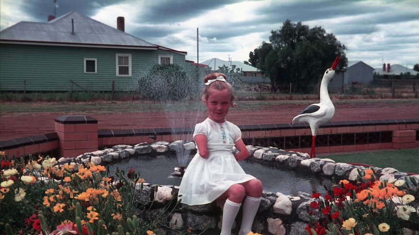 Lisa McManus, as a young girl wearing a dress, sits on the edge of a fountain in a garden.