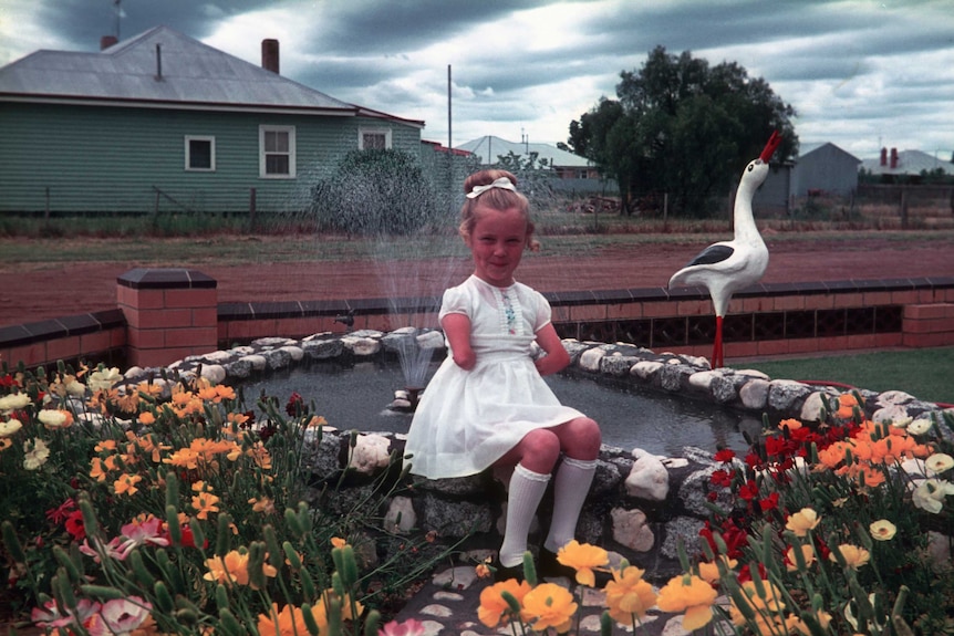 Lisa McManus, as a young girl wearing a dress, sits on the edge of a fountain in a garden.