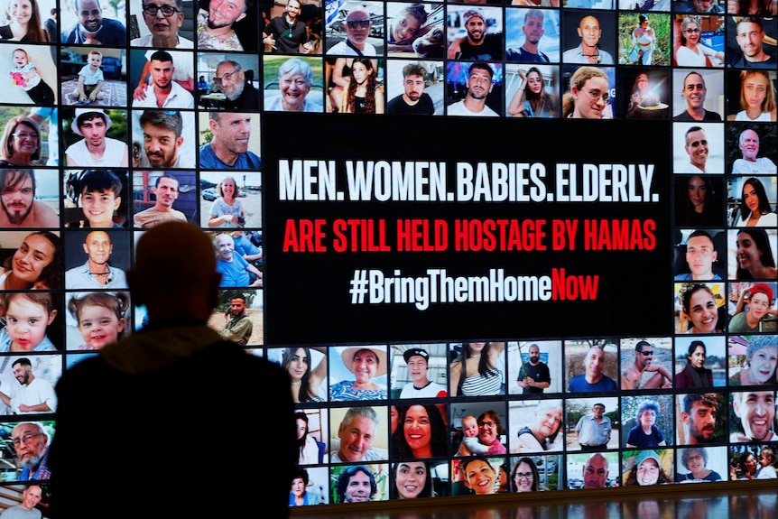 A silhouette of a person is seen in front of a display showing rows of photos of hostages on a wall.