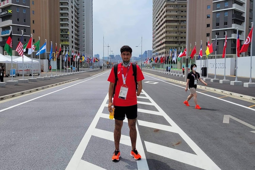 A middle eastern man in red with partially amputated left arm stands in middle of empty road flanked by flags on a sunny day.