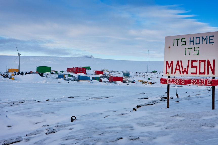 A sign reads "It's Home, It's Mawson" on a field of ice in front of an Antarctic station.