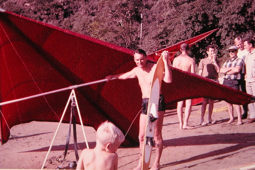 An old colour photo of a man in swimmers holding a red hang glider on the sand.