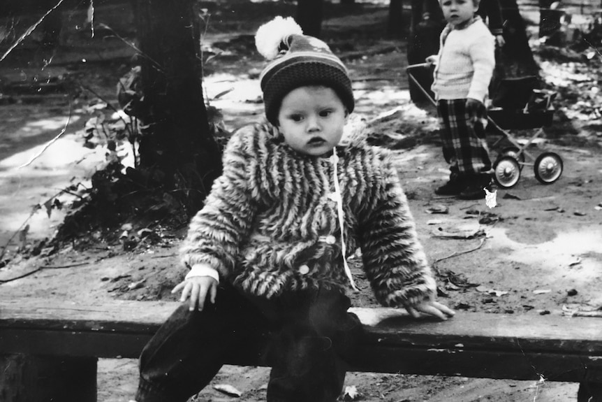 Old black and white photo of a baby sitting on a bench wearing a beanie and coat.