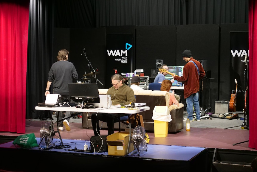 A guitarist in a red jumper stands with his back to the camera alongside other musicians in a recording studio.