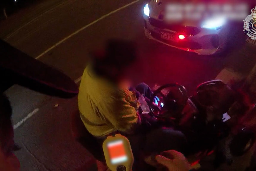 A man in high-vis sits on a ride-on lawnmower at night. He is being stopped by police.