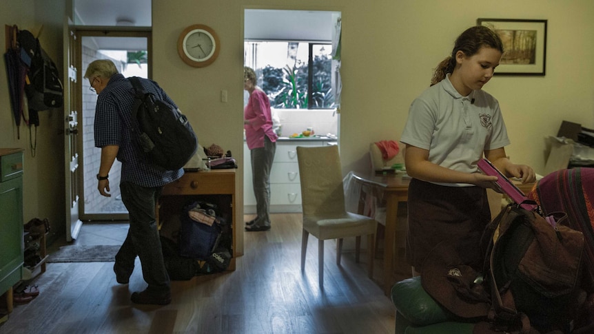 A man walking inside, as a woman washes dishes and a young girl packs a school bag