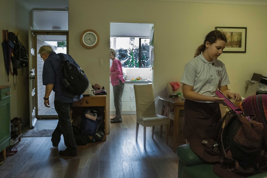 A man walking inside, as a woman washes dishes and a young girl packs a school bag