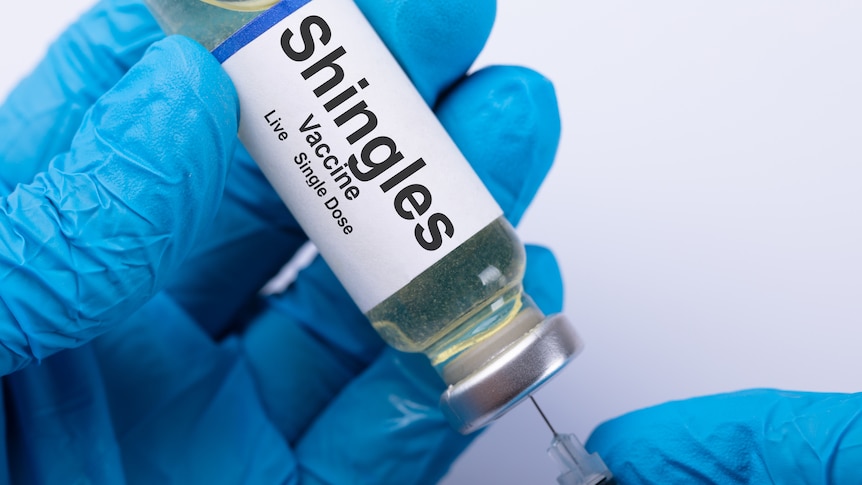 syringe with 'shingles vaccine' on the label held by two blue gloves