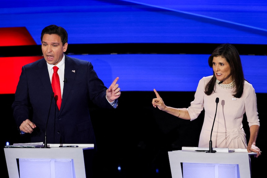 A man and a woman hold out pointed fingers at each other while standing on a debate stage.