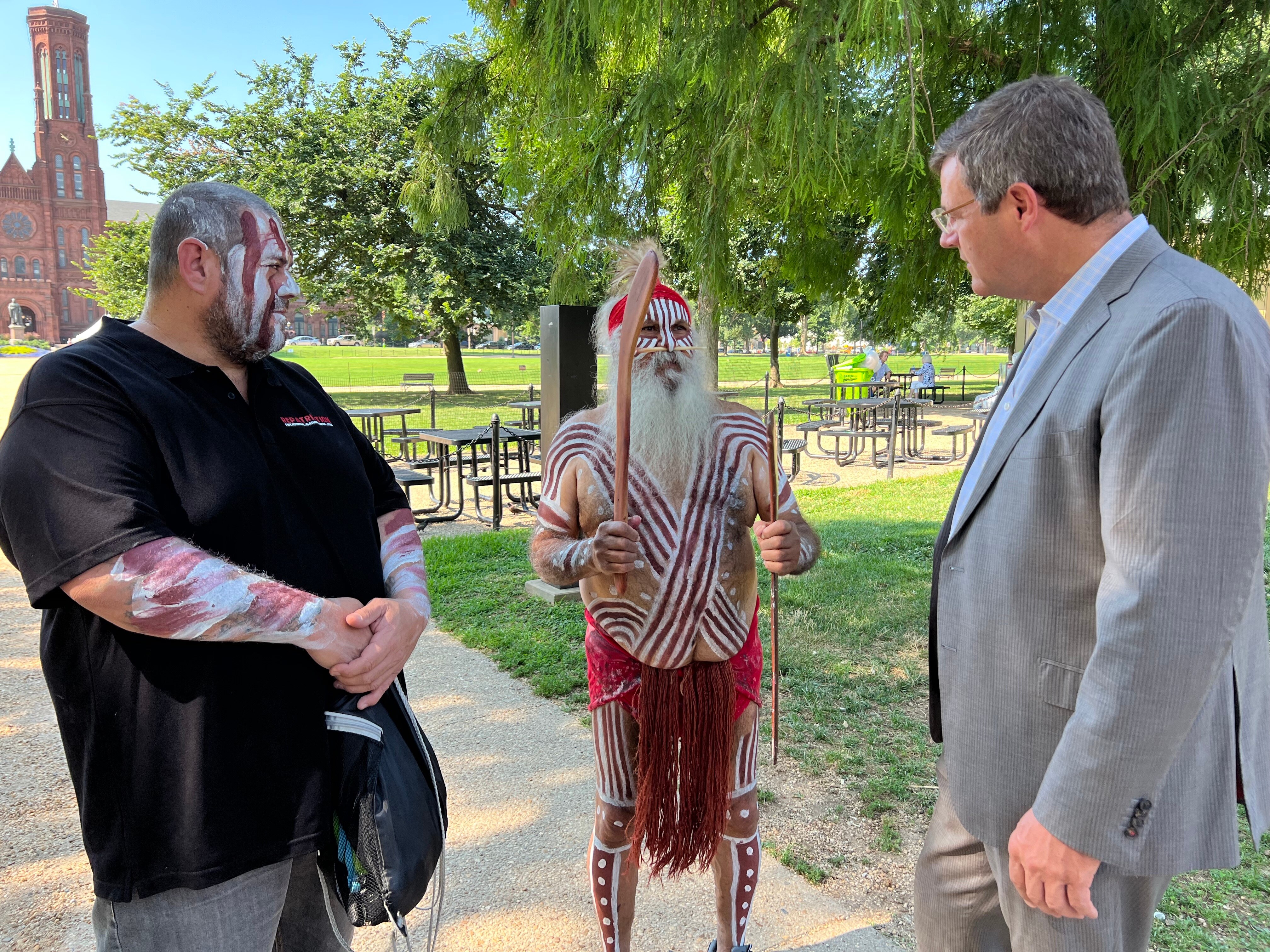 Two Indigenous men, one in a black polo shirt and the other in traditional dress, stand next to a white an in a suit