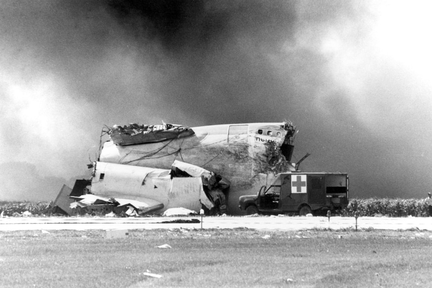 An ambulance parks near mangled parts of a large passenger airplane while smoke fills the air.