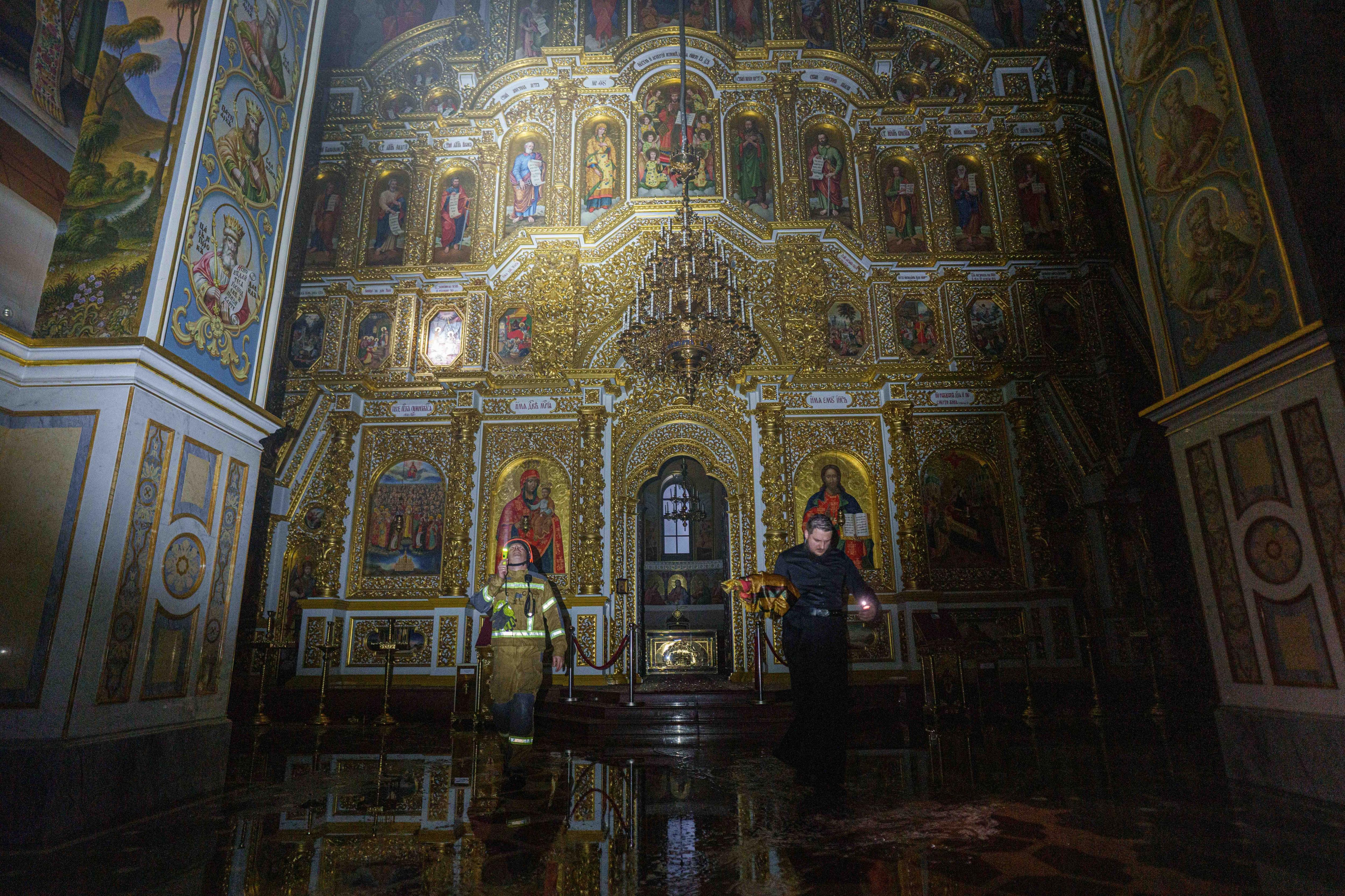 A priest carries church inventory out of a dark Cathedral after a fire. The walls are lined with gold-covered religious symbols.