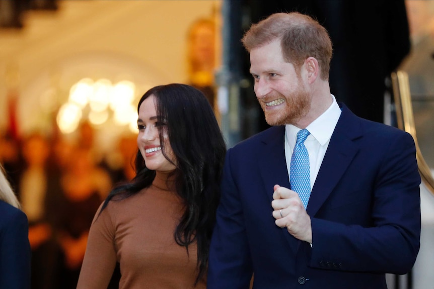 Prince Harry and Meghan, Duchess of Sussex leave a building. Both are smiling excitedly.