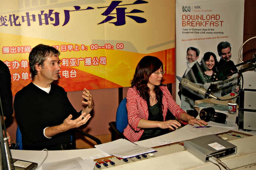 Man and woman sitting next to each other in front of a radio microphone with Chinese language and ABC posters behind them.