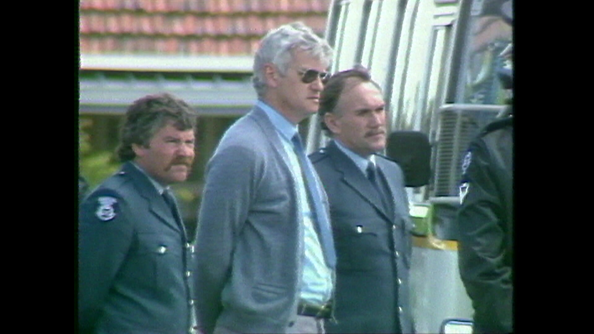 Un hombre de pelo gris lleva gafas de sol y corbata. Se encuentra junto a dos policías y un minibús.
