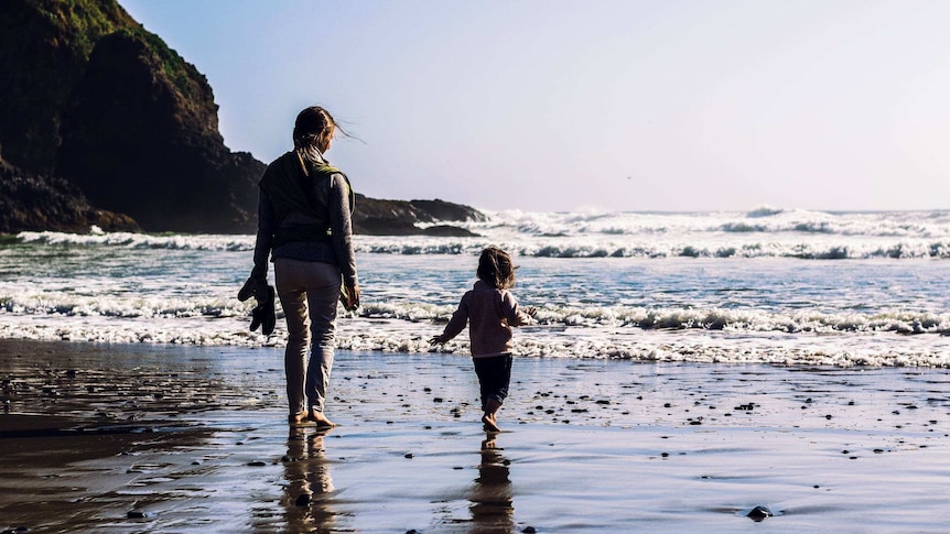 A woman faces the ocean with her young child standing next to her, as the waves come crashing in to the shore. The sky is blue.