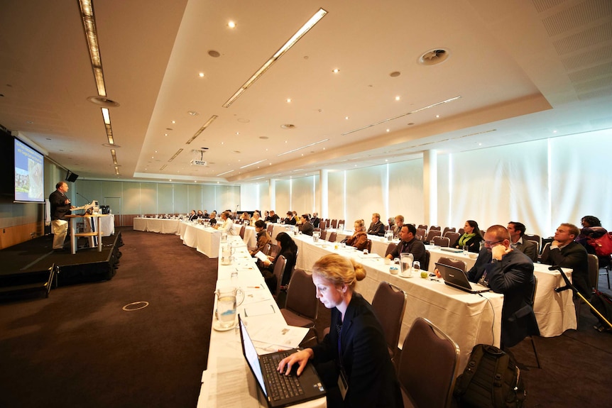 Around 30 delegates sit at long tables in a large room listening to a speaker at the Perth Conference and Exhibition Centre.