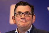 Daniel Andrews, in a suit, speaks to reporters in front of a purple health department backdrop.