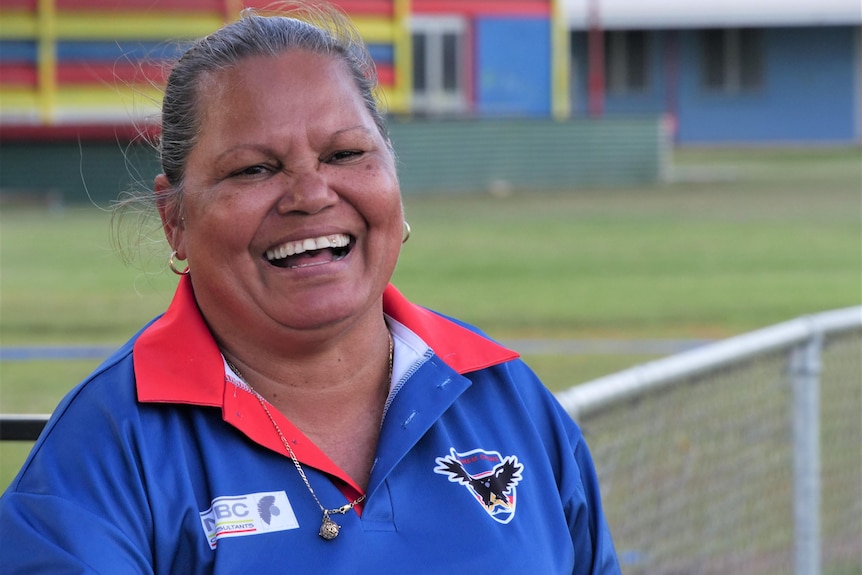 A woman smiles standing on the edge of a football oval in a remote community. 