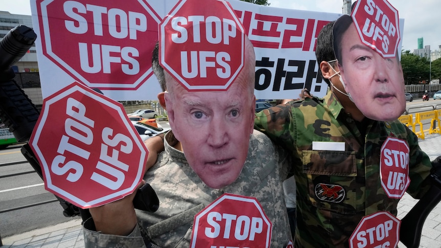 Two people in military gear wear masks of the US and Korean presidents while holding "STOP UFS" signs 
