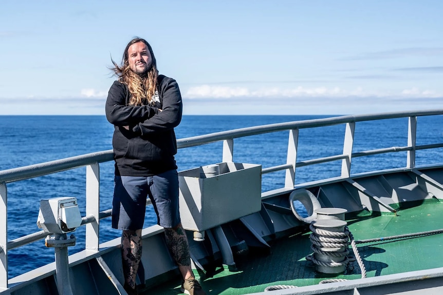 A man with long brain hair and wearing a black hoodie and shorts stands on a boat with arms crossed. 