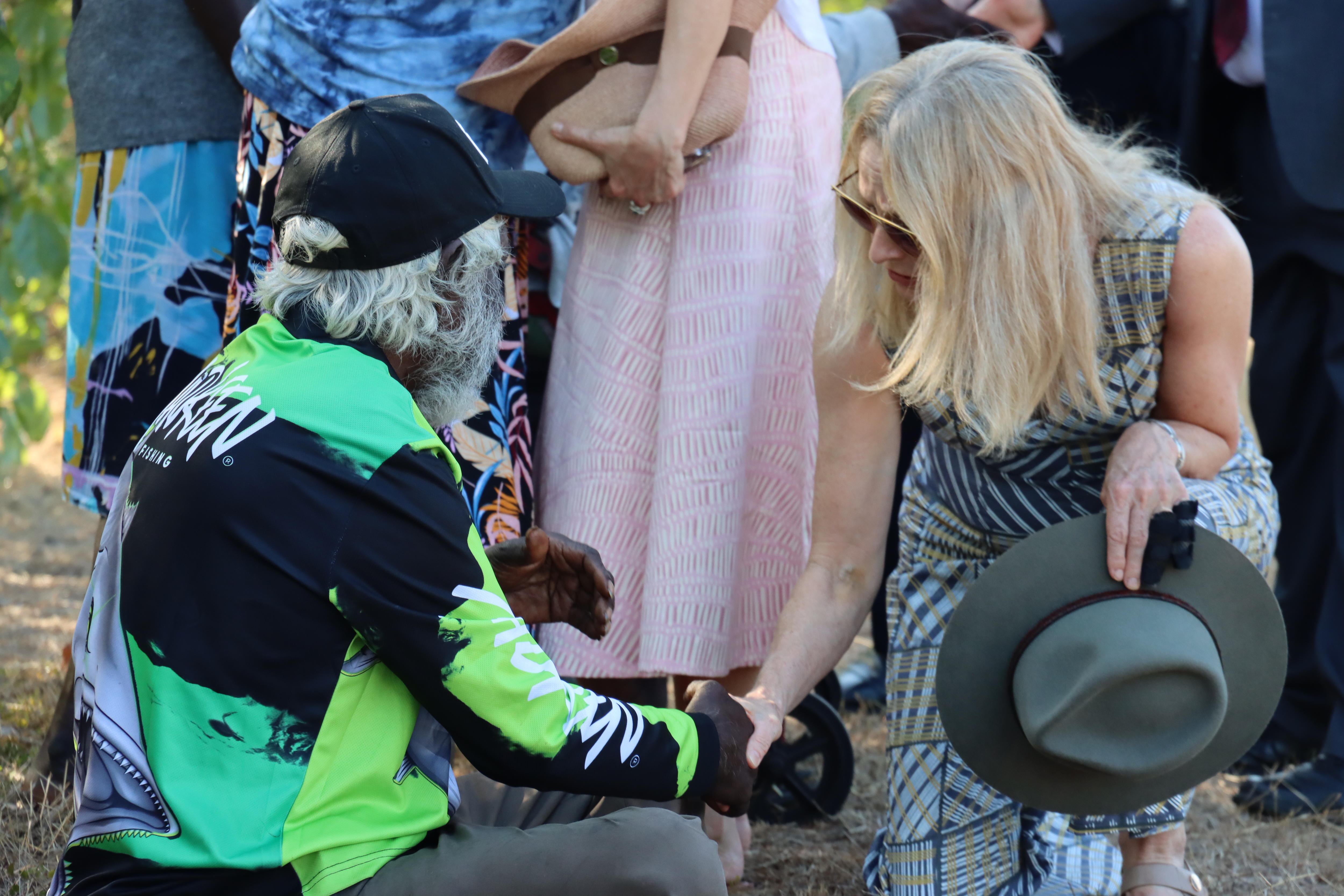 A blonde woman holding her hat shakes the hand of an older man with white hair. Behind them are many other people.