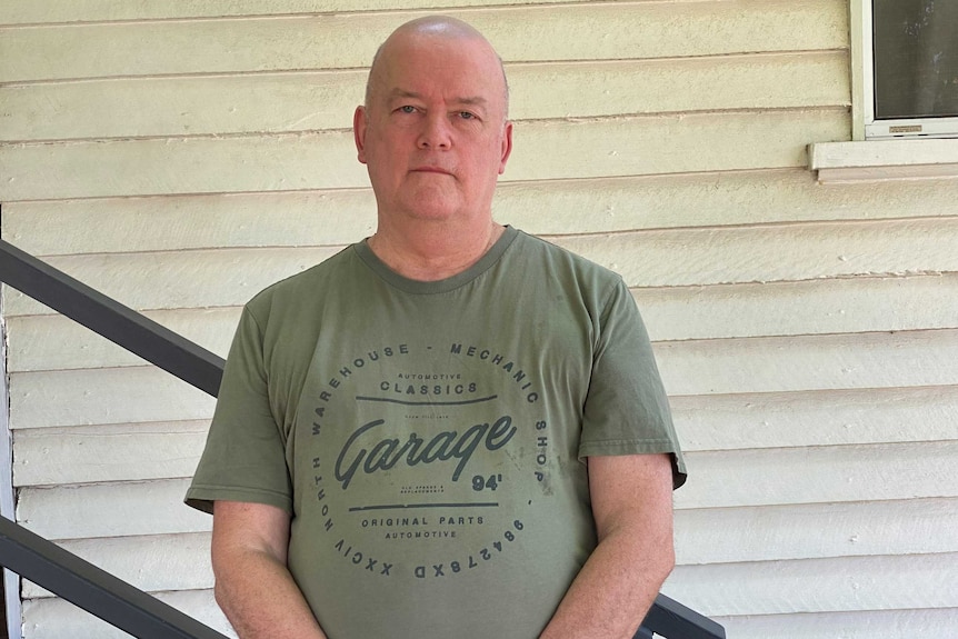 A slightly overweight bald man in green T-shirt in his 50s stands by stairs in front of small house.