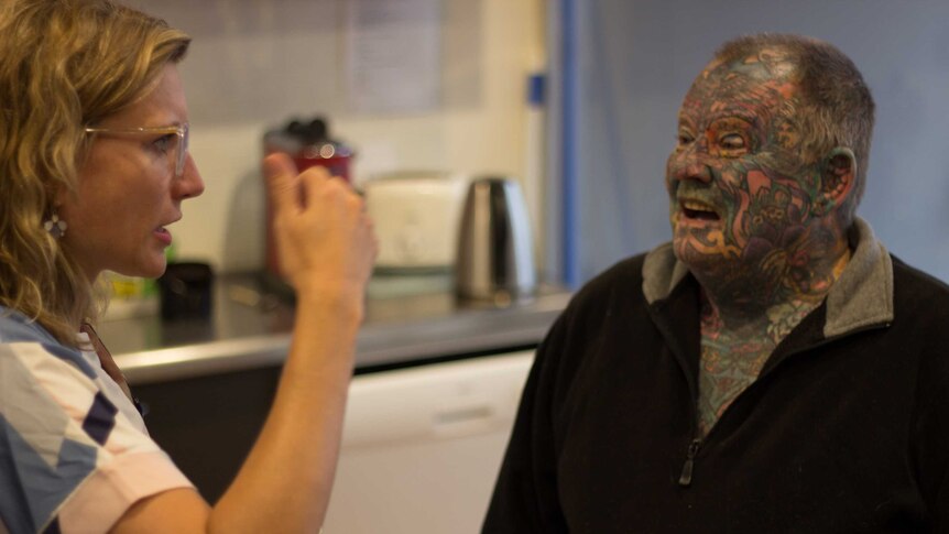 A woman with glasses speaks to a man with a heavily tattooed face in a kitchen.