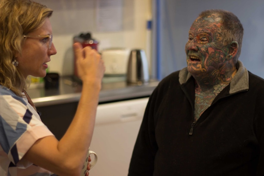 A woman with glasses speaks to a man with a heavily tattooed face in a kitchen.