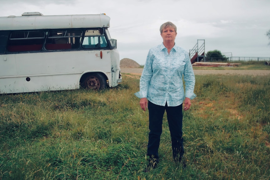 A middle-aged woman with short hair stands on a rural property with an old bus in the background.