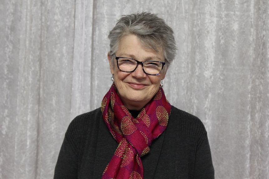 A woman with grey hair and a pink scarf and glasses smiles at the camera 