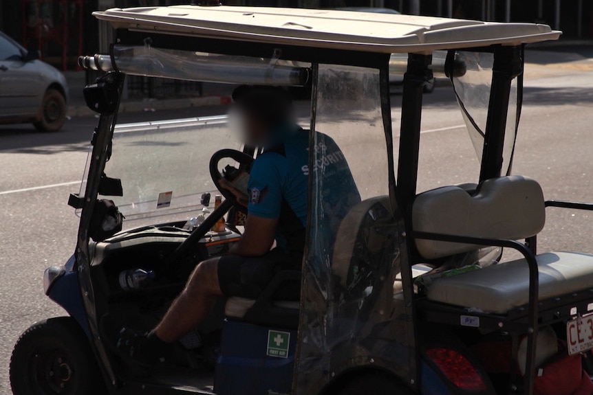 A man in a blue shirt sits in a golf buggy looking out at the city street in front of him. 