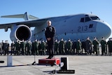 A man stands on a podium speaking into a microphone with a miltary transport jet as a backdrop