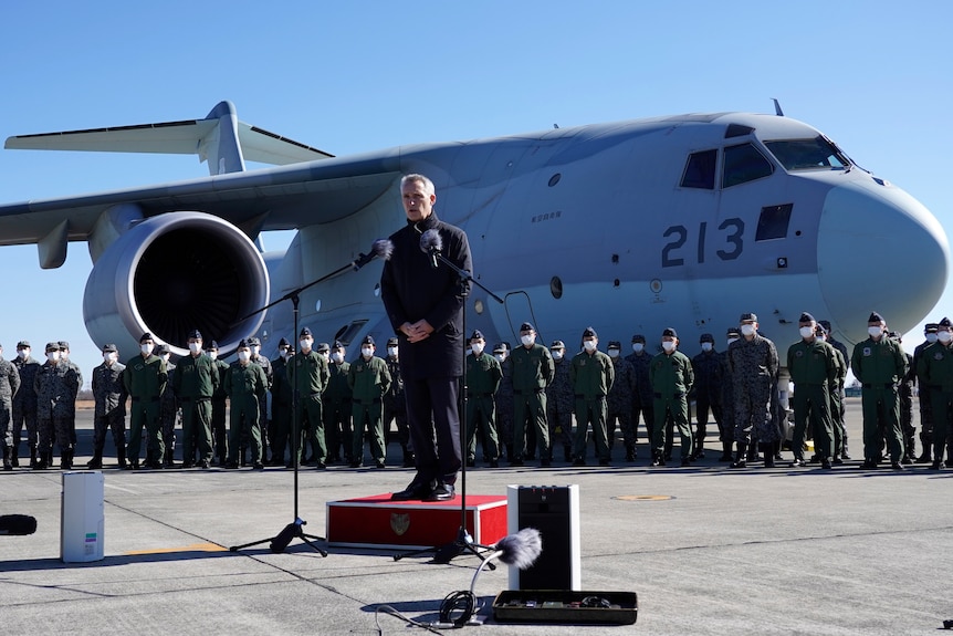 A man stands on a podium speaking into a microphone with a miltary transport jet as a backdrop