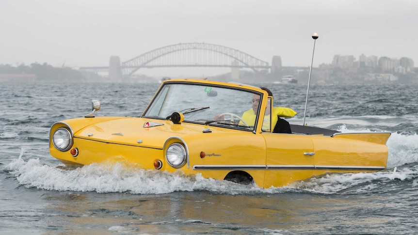 A yellow amphibious car floats on Sydney Harbour with the Sydney Harbour Bridge seen in the distance behind.