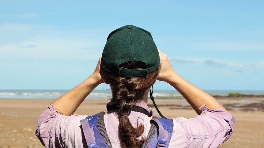 A girl holding binoculars and looking to the horizon, photographed from behind.