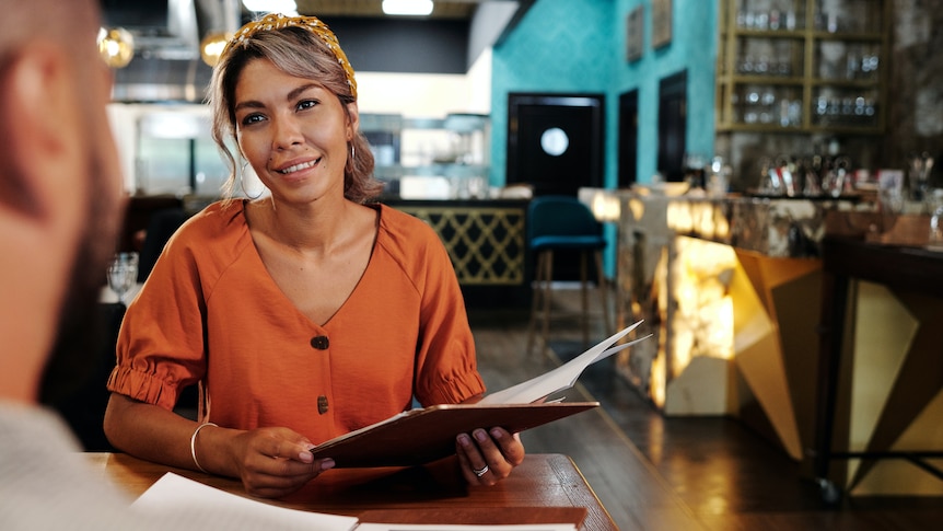 A woman wearing an orange dress holds a menu and looks at a man on a date in a resturant.