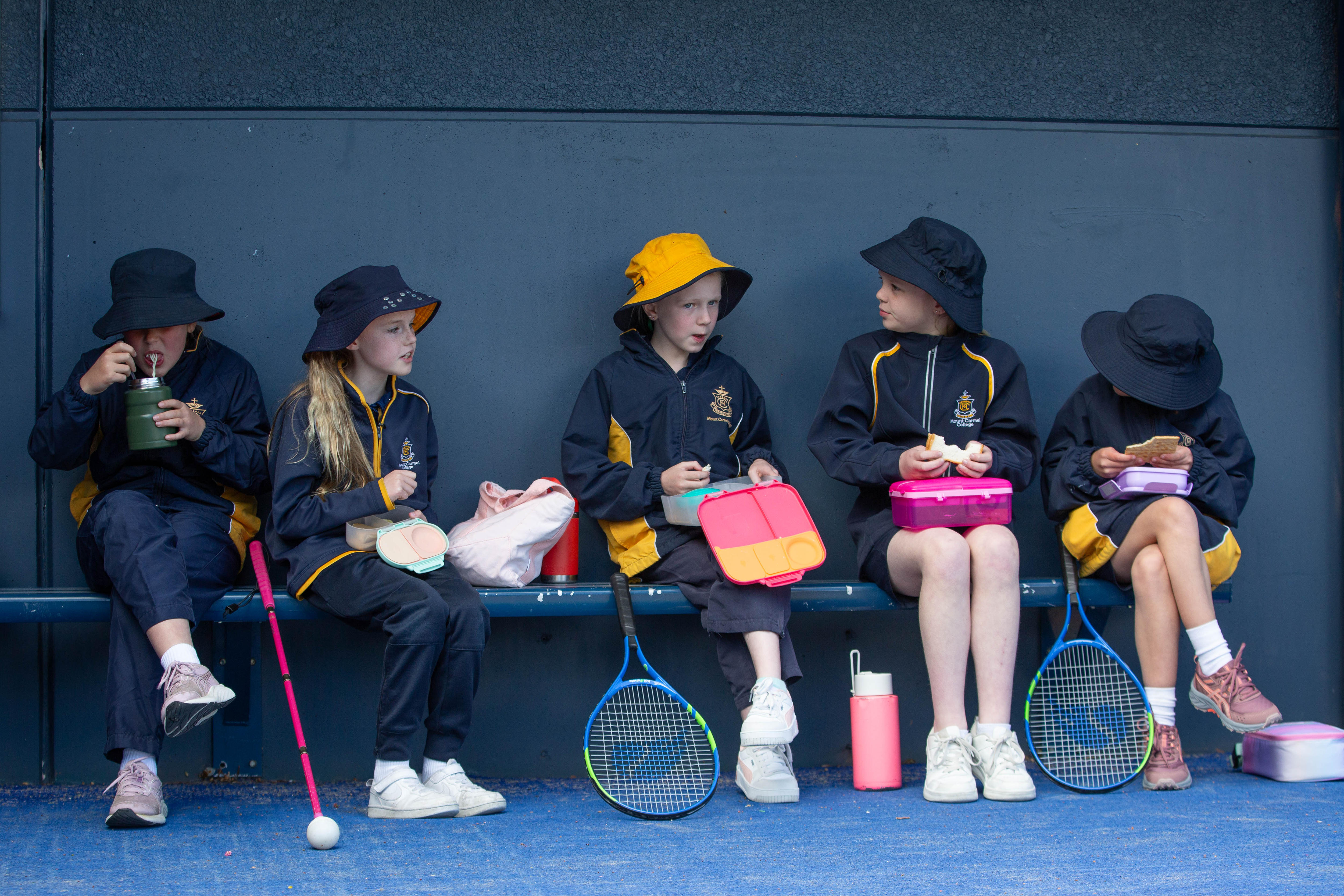 Cinco colegialas se sientan en un banco y almuerzan. Dos tienen raquetas de tenis. Uno tiene un palo.
