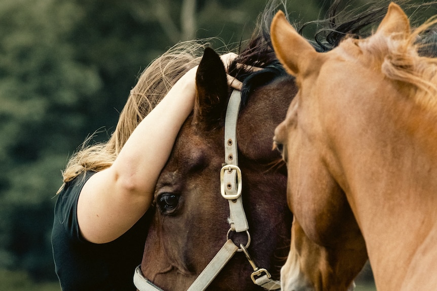 Amy, who has blonde hair, hides her face in the mane of a horse outside.