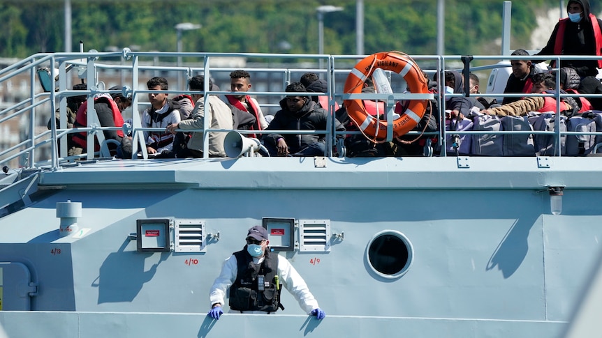 A group of people thought to be migrants are brought into a dock on a large grey vessel.