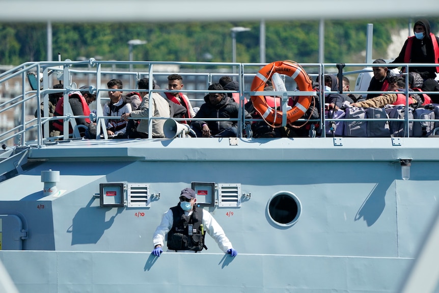 Un groupe de personnes considérées comme des migrants est amené à quai sur un grand navire gris.