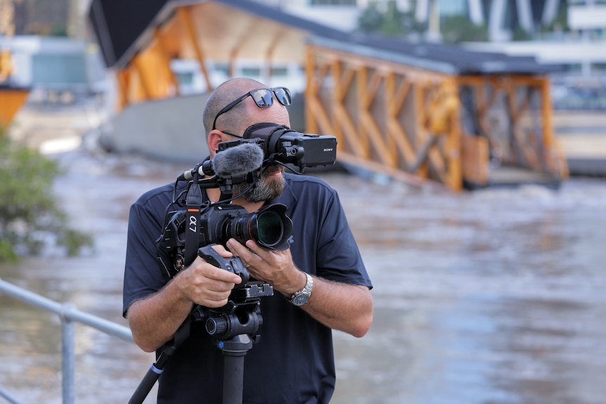 Cameraman looking through camera lens with backdrop of flooded river.