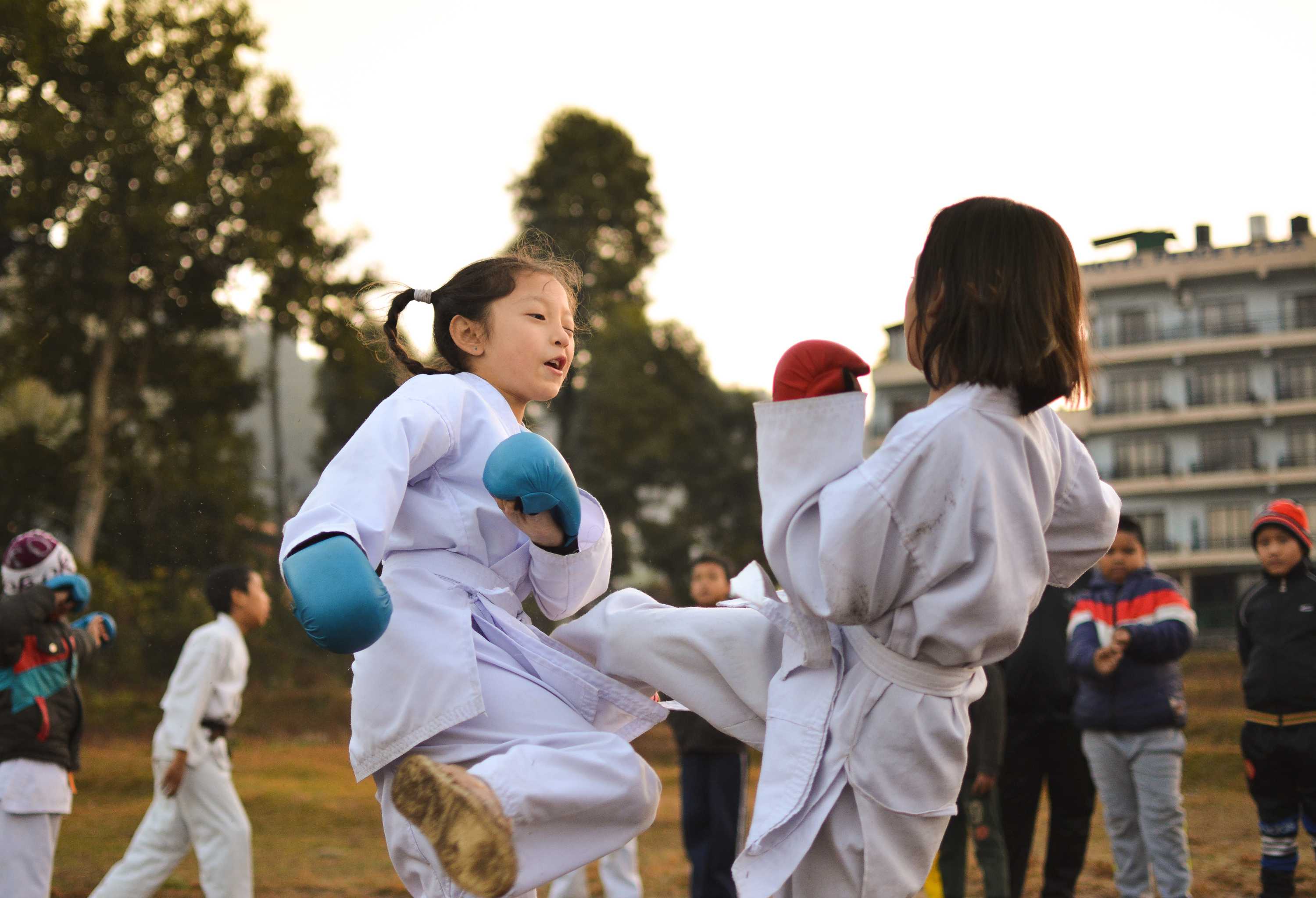 Two young children in martial arts uniforms spar outdoors, surrounded by other kids