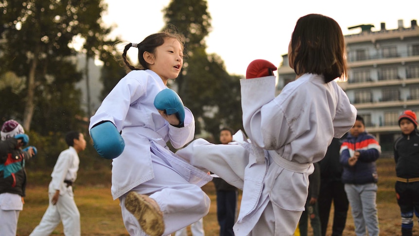 Two young children in martial arts uniforms spar outdoors, surrounded by other kids